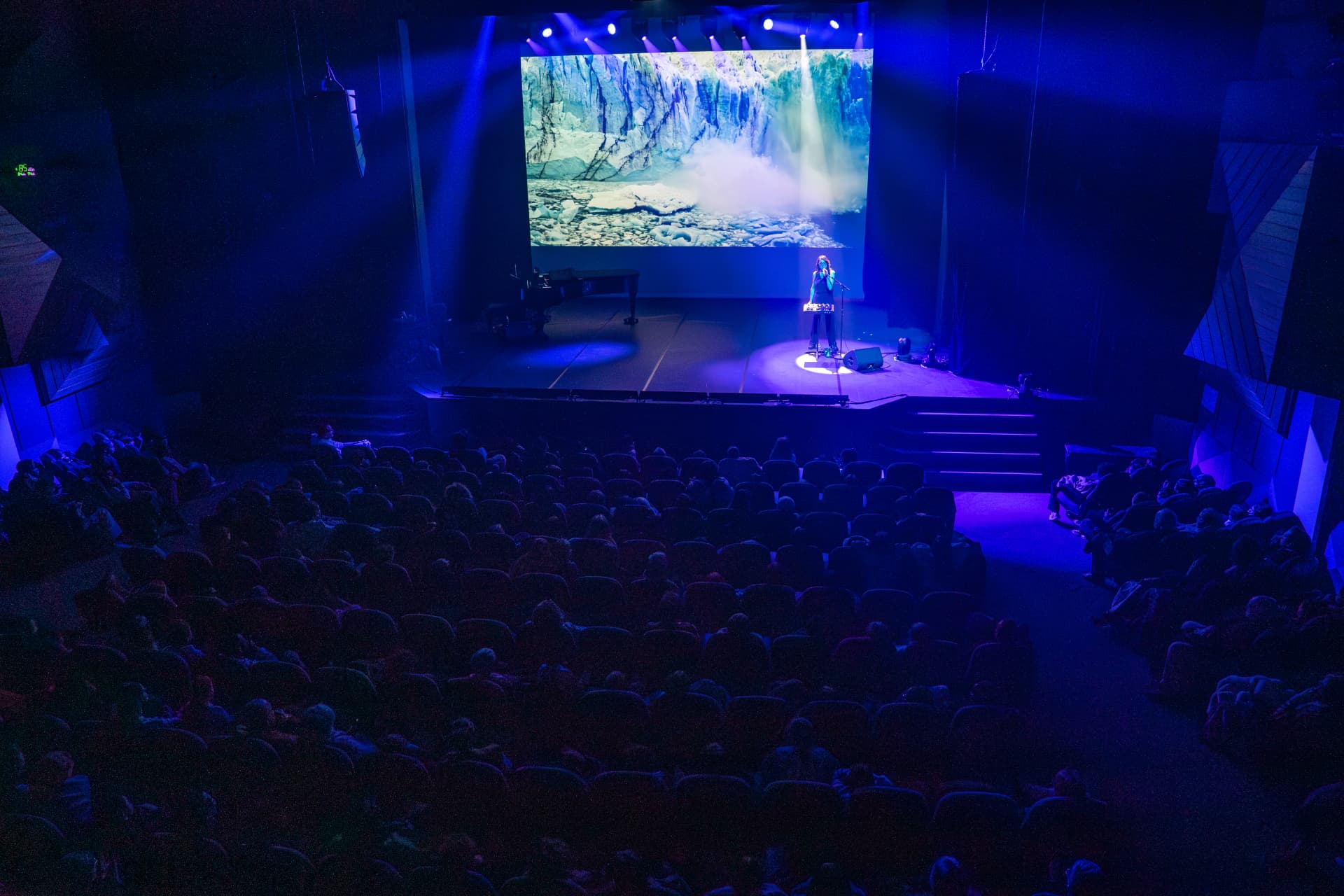 Vue de la salle plongée dans un bleu profond pendant Résonances arctiques : une artiste sur scène sous un faisceau de lumière, un glacier projeté sur grand écran